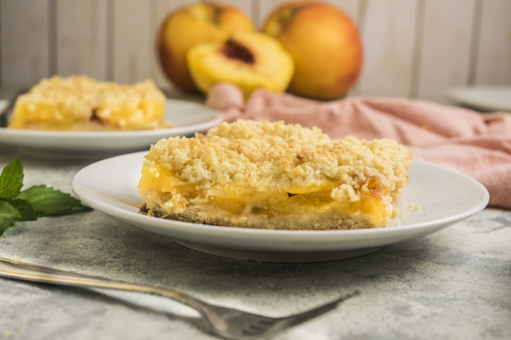 A slice of sbriciolata di pesche, an Italian peach crumble cake, served on a white plate with soft-focus peaches and a second slice in the background.